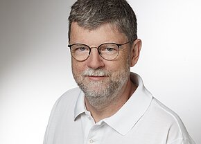 Porträt eines Mannes mit grauen Haaren und Brille. Er trägt ein weißes Polo-Shirt und hat einen freundlichen Gesichtsausdruck. Der Hintergrund ist hell und neutral.